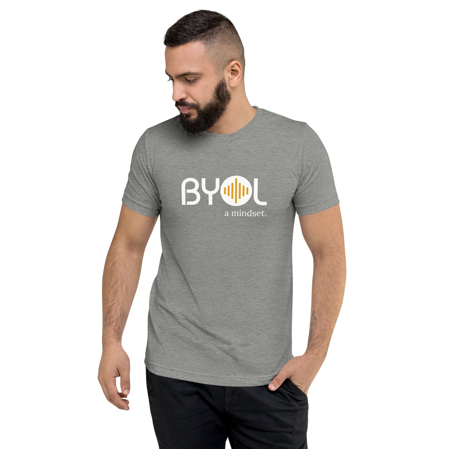 A young man with short dark hair and a beard wearing a gray "BYOL: a mindset" T-shirt, standing with one hand in his pocket and looking down. The T-shirt features the "BYOL" logo in white and yellow on the front and is available in multiple colors (black, red, gray, pink, gold, plus more) and sizes (S-3XL). The "be you out loud" logo is displayed at the top.