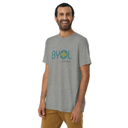 A young man with short dark hair and a beard wearing a tri-blend grey "BYOL: a mindset" T-shirt, smiling and standing with his arms at his sides. The T-shirt features the "BYOL" logo in teal and is available in multiple colors (black, red, gray, pink, yellow, plus more) and sizes (S-3XL). The "be you out loud" logo is displayed at the top.