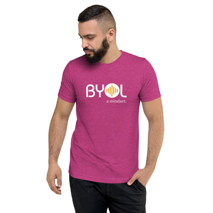 A young man with short dark hair and a beard wearing a berry tri-blend "BYOL: a mindset" T-shirt, standing with one hand in his pocket and looking down. The T-shirt features the "BYOL" logo in white and yellow on the front and is available in multiple colors (black, red, gray, pink, gold, plus more) and sizes (S-3XL). The "be you out loud" logo is displayed at the top.