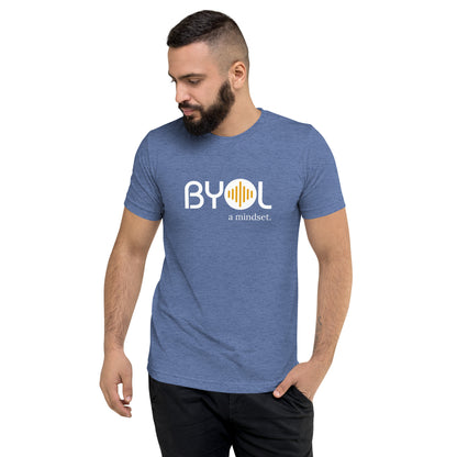 A young man with short dark hair and a beard wearing a royal blue tri-blend "BYOL: a mindset" T-shirt, standing with one hand in his pocket and looking down. The T-shirt features the "BYOL" logo in white and yellow on the front and is available in multiple colors (black, red, gray, pink, gold, plus more) and sizes (S-3XL). The "be you out loud" logo is displayed at the top.