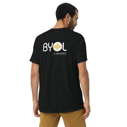 A young man with short dark hair wearing a solid black tri-blend "BYOL: a mindset" T-shirt, viewed from the back. The T-shirt features the "BYOL" logo in white and yellow on the back and is available in multiple colors (black, red, gray, pink, beige, plus more) and sizes (S-3XL). The "be you out loud" logo is displayed at the top.