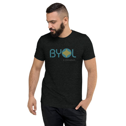 A young man with short dark hair and a beard wearing a charcoal grey "BYOL: a mindset" T-shirt, standing with one hand in his pocket and looking down. The T-shirt features the "BYOL" logo in blue and yellow on the front and is available in multiple colors (black, red, gray, pink, gold, plus more) and sizes (S-3XL). The "be you out loud" logo is displayed at the top.