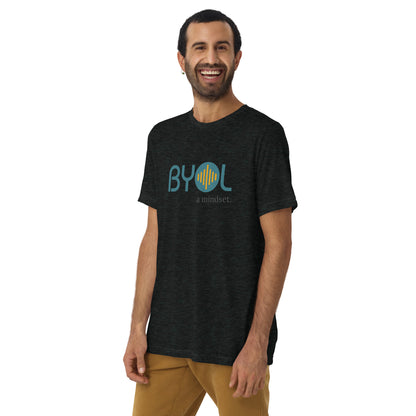 A young man with short dark hair and a beard wearing a charcoal tri-blend "BYOL: a mindset" T-shirt, smiling and standing with his arms at his sides. The T-shirt features the "BYOL" logo in teal and is available in multiple colors (black, red, gray, pink, yellow, plus more) and sizes (S-3XL). The "be you out loud" logo is displayed at the top.