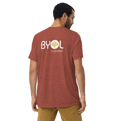 A young man with short dark hair wearing a clay tri-blend "BYOL: a mindset" T-shirt, viewed from the back. The T-shirt features the "BYOL" logo in white and yellow on the back and is available in multiple colors (black, red, gray, pink, beige, plus more) and sizes (S-3XL). The "be you out loud" logo is displayed at the top.