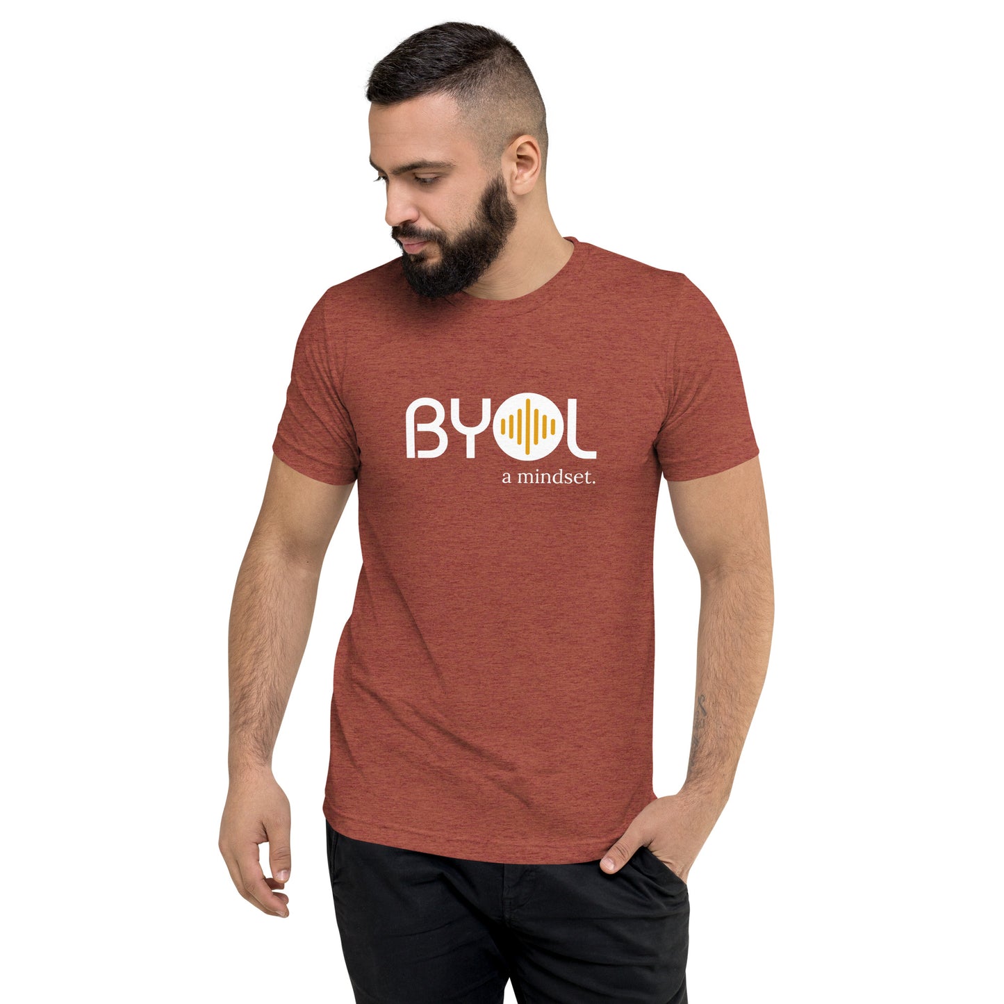 A young man with short dark hair and a beard wearing a clay trei-blend "BYOL: a mindset" T-shirt, standing with one hand in his pocket and looking down. The T-shirt features the "BYOL" logo in white and yellow on the front and is available in multiple colors (black, red, gray, pink, gold, plus more) and sizes (S-3XL). The "be you out loud" logo is displayed at the top.