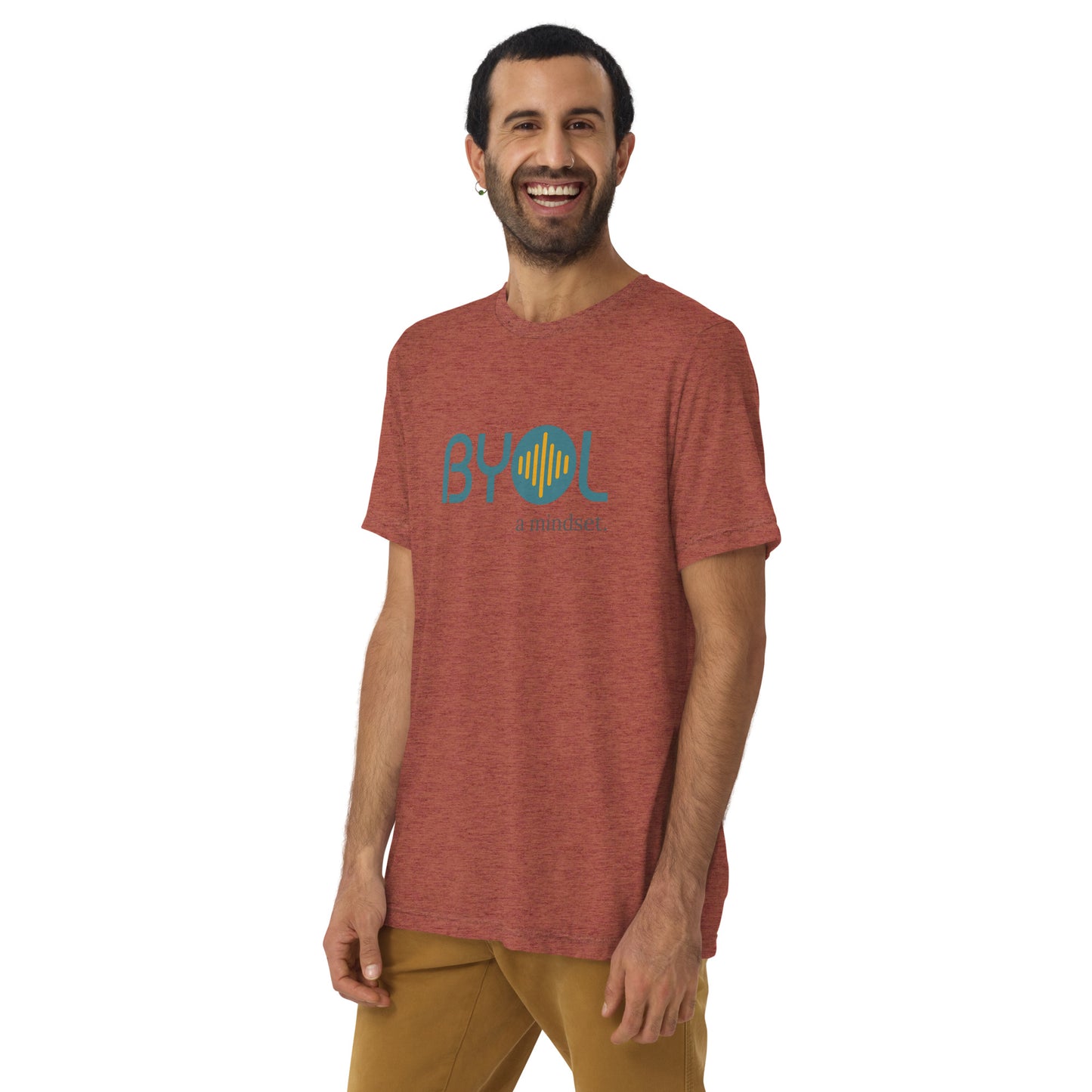 A young man with short dark hair and a beard wearing a mauve tri-blend "BYOL: a mindset" T-shirt, smiling and standing with his arms at his sides. The T-shirt features the "BYOL" logo in teal and is available in multiple colors (black, red, gray, pink, yellow, plus more) and sizes (S-3XL). The "be you out loud" logo is displayed at the top.