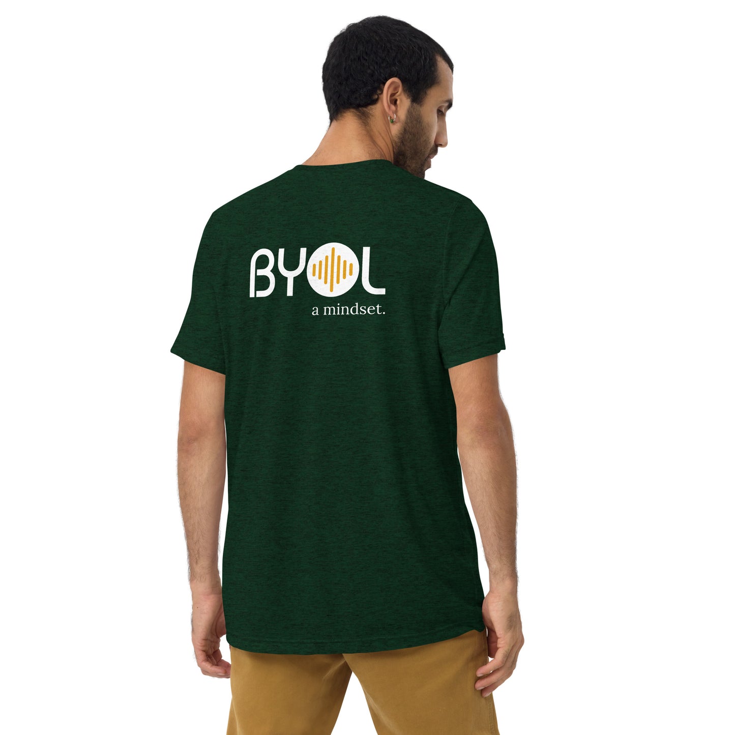 A young man with short dark hair wearing a emerald green tri-blend "BYOL: a mindset" T-shirt, viewed from the back. The T-shirt features the "BYOL" logo in white and yellow on the back and is available in multiple colors (black, red, gray, pink, beige, plus more) and sizes (S-3XL). The "be you out loud" logo is displayed at the top.