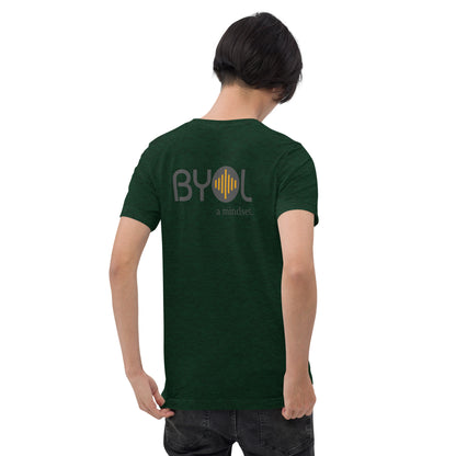 A young man with short black hair wearing a emerald green tri-blend "BYOL: a mindset" T-shirt, viewed from the back. The T-shirt features the "BYOL" logo in black and yellow on the back and is available in multiple colors (black, red, gray, pink, gold, plus more) and sizes (S-3XL). The "be you out loud" logo is displayed at the top.