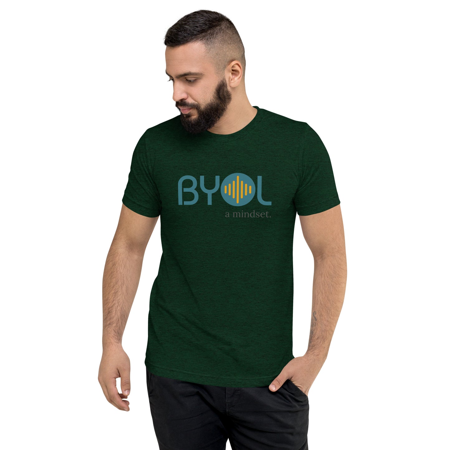 A young man with short dark hair and a beard wearing a emerald tri-blenc "BYOL: a mindset" T-shirt, standing with one hand in his pocket and looking down. The T-shirt features the "BYOL" logo in blue and yellow on the front and is available in multiple colors (black, red, gray, pink, gold, plus more) and sizes (S-3XL). The "be you out loud" logo is displayed at the top.