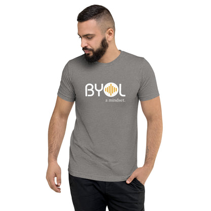 A young man with short dark hair and a beard wearing a gray "BYOL: a mindset" T-shirt, standing with one hand in his pocket and looking down. The T-shirt features the "BYOL" logo in white and yellow on the front and is available in multiple colors (black, red, gray, pink, gold, plus more) and sizes (S-3XL). The "be you out loud" logo is displayed at the top.