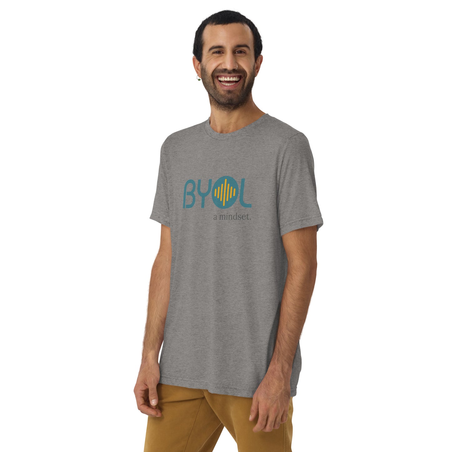 A young man with short dark hair and a beard wearing a grey tri-blend "BYOL: a mindset" T-shirt, smiling and standing with his arms at his sides. The T-shirt features the "BYOL" logo in teal and is available in multiple colors (black, red, gray, pink, yellow, plus more) and sizes (S-3XL). The "be you out loud" logo is displayed at the top.