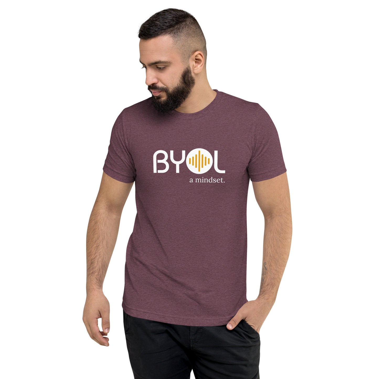 A young man with short dark hair and a beard wearing a maroon tri-blend "BYOL: a mindset" T-shirt, standing with one hand in his pocket and looking down. The T-shirt features the "BYOL" logo in white and yellow on the front and is available in multiple colors (black, red, gray, pink, gold, plus more) and sizes (S-3XL). The "be you out loud" logo is displayed at the top.