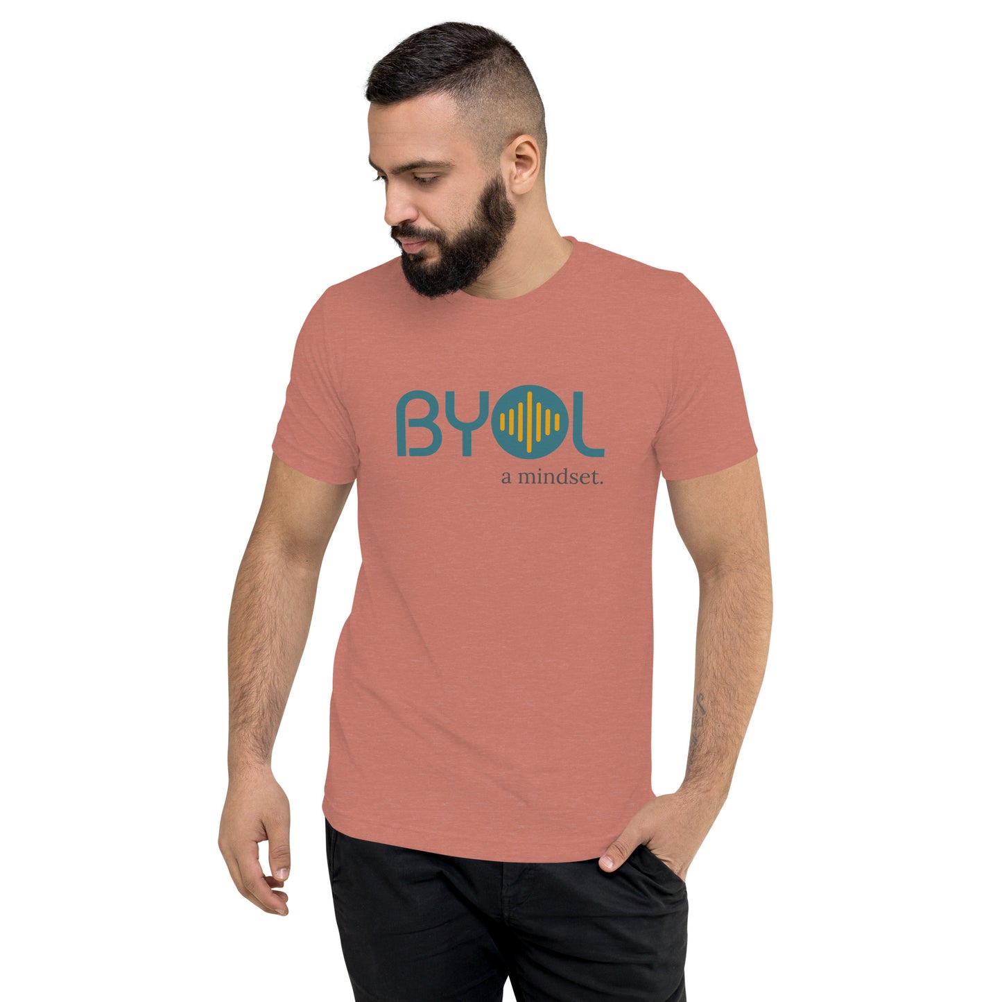 A young man with short dark hair and a beard wearing a mauve tri-blend "BYOL: a mindset" T-shirt, standing with one hand in his pocket and looking down. The T-shirt features the "BYOL" logo in blue and yellow on the front and is available in multiple colors (black, red, gray, pink, gold, plus more) and sizes (S-3XL). The "be you out loud" logo is displayed at the top.