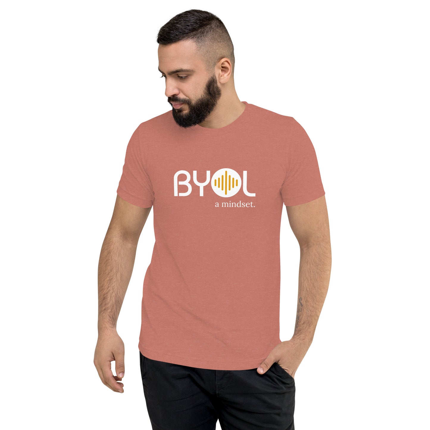 A young man with short dark hair and a beard wearing a mauve tri-blend "BYOL: a mindset" T-shirt, standing with one hand in his pocket and looking down. The T-shirt features the "BYOL" logo in white and yellow on the front and is available in multiple colors (black, red, gray, pink, gold, plus more) and sizes (S-3XL). The "be you out loud" logo is displayed at the top.