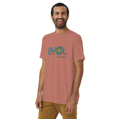 A young man with short dark hair and a beard wearing a red tri-blend "BYOL: a mindset" T-shirt, smiling and standing with his arms at his sides. The T-shirt features the "BYOL" logo in teal and is available in multiple colors (black, red, gray, pink, yellow, plus more) and sizes (S-3XL). The "be you out loud" logo is displayed at the top.