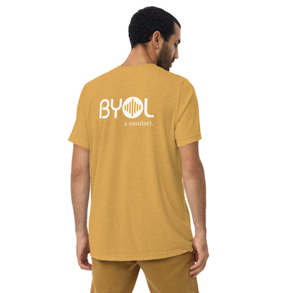 A young man with short dark hair wearing a mustard tri-blend "BYOL: a mindset" T-shirt, viewed from the back. The T-shirt features the "BYOL" logo in white and yellow on the back and is available in multiple colors (black, red, gray, pink, beige, plus more) and sizes (S-3XL). The "be you out loud" logo is displayed at the top.