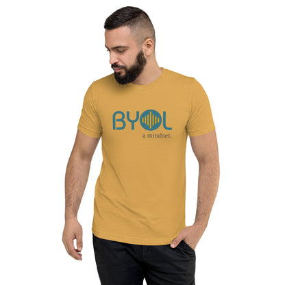 A young man with short dark hair and a beard wearing a mustard tri-blend "BYOL: a mindset" T-shirt, standing with one hand in his pocket and looking down. The T-shirt features the "BYOL" logo in blue and yellow on the front and is available in multiple colors (black, red, gray, pink, gold, plus more) and sizes (S-3XL). The "be you out loud" logo is displayed at the top.