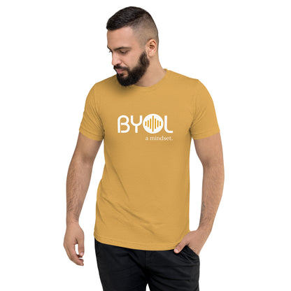 A young man with short dark hair and a beard wearing a mustard yellow tri-blend "BYOL: a mindset" T-shirt, standing with one hand in his pocket and looking down. The T-shirt features the "BYOL" logo in white and yellow on the front and is available in multiple colors (black, red, gray, pink, gold, plus more) and sizes (S-3XL). The "be you out loud" logo is displayed at the top.