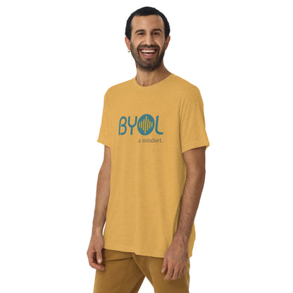 A young man with short dark hair and a beard wearing a mustard tri-blend "BYOL: a mindset" T-shirt, smiling and standing with his arms at his sides. The T-shirt features the "BYOL" logo in teal and is available in multiple colors (black, red, gray, pink, yellow, plus more) and sizes (S-3XL). The "be you out loud" logo is displayed at the top.