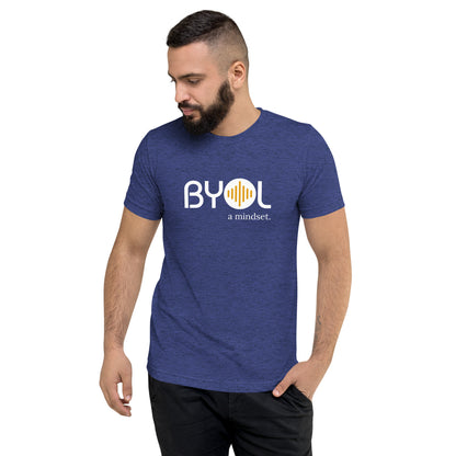 A young man with short dark hair and a beard wearing a navy blue tri-blend "BYOL: a mindset" T-shirt, standing with one hand in his pocket and looking down. The T-shirt features the "BYOL" logo in white and yellow on the front and is available in multiple colors (black, red, gray, pink, gold, plus more) and sizes (S-3XL). The "be you out loud" logo is displayed at the top.