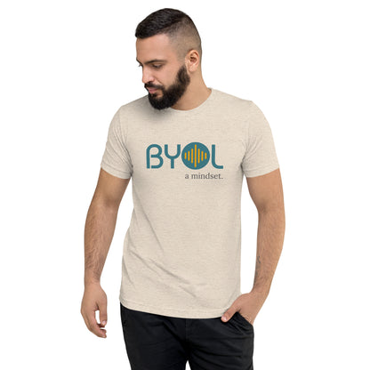 A young man with short dark hair and a beard wearing a oatmeal tri-blend "BYOL: a mindset" T-shirt, standing with one hand in his pocket and looking down. The T-shirt features the "BYOL" logo in blue and yellow on the front and is available in multiple colors (black, red, gray, pink, gold, plus more) and sizes (S-3XL). The "be you out loud" logo is displayed at the top.