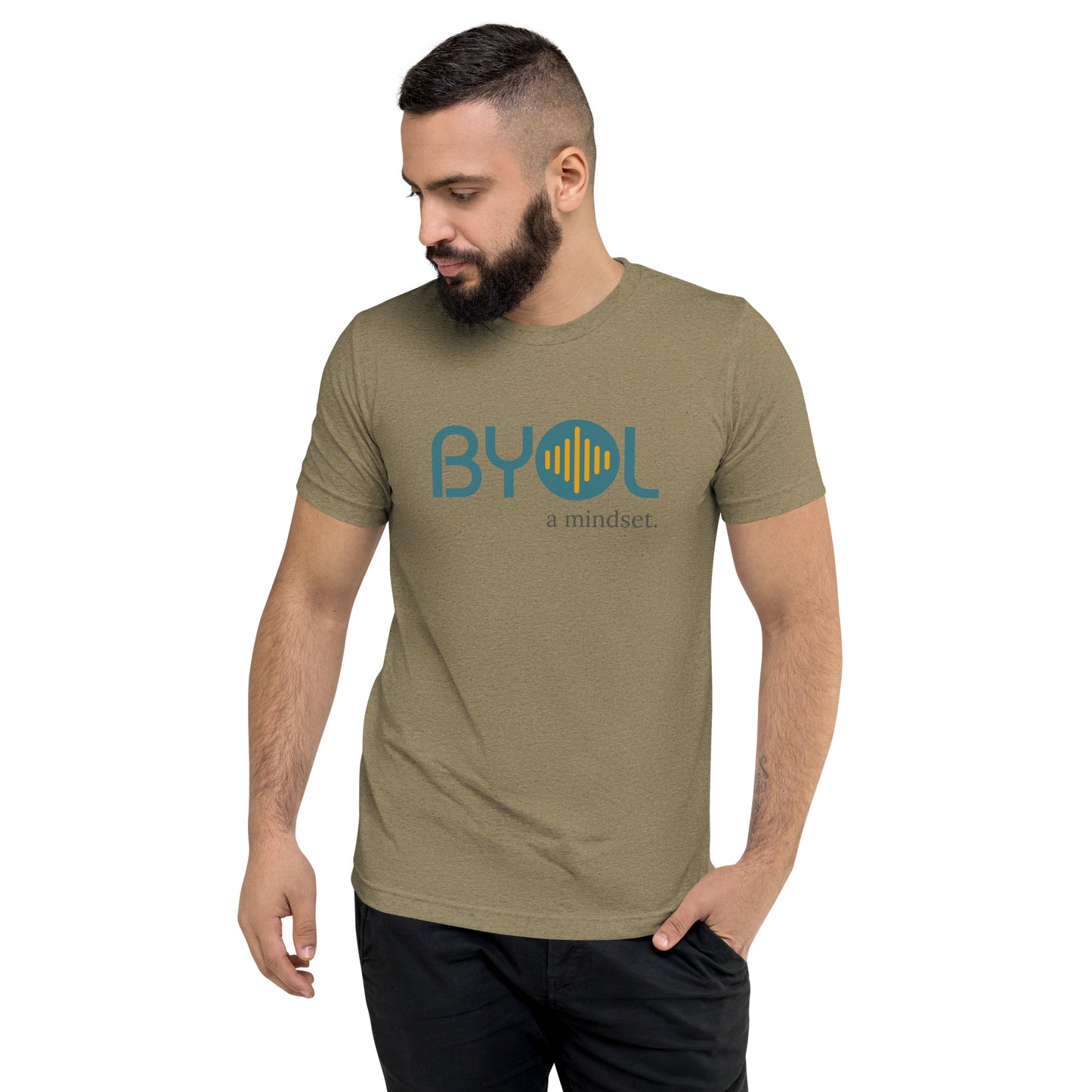 A young man with short dark hair and a beard wearing a olive tri-blend "BYOL: a mindset" T-shirt, standing with one hand in his pocket and looking down. The T-shirt features the "BYOL" logo in blue and yellow on the front and is available in multiple colors (black, red, gray, pink, gold, plus more) and sizes (S-3XL). The "be you out loud" logo is displayed at the top.