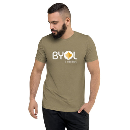 A young man with short dark hair and a beard wearing a olive green tri-blend "BYOL: a mindset" T-shirt, standing with one hand in his pocket and looking down. The T-shirt features the "BYOL" logo in white and yellow on the front and is available in multiple colors (black, red, gray, pink, gold, plus more) and sizes (S-3XL). The "be you out loud" logo is displayed at the top.