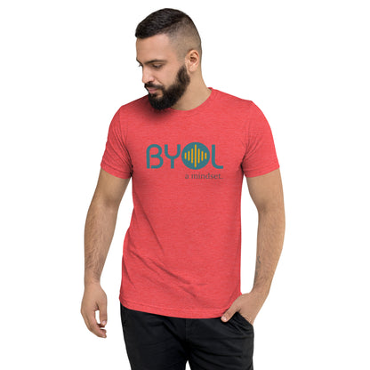 A young man with short dark hair and a beard wearing a red tri-blend "BYOL: a mindset" T-shirt, standing with one hand in his pocket and looking down. The T-shirt features the "BYOL" logo in blue and yellow on the front and is available in multiple colors (black, red, gray, pink, gold, plus more) and sizes (S-3XL). The "be you out loud" logo is displayed at the top.