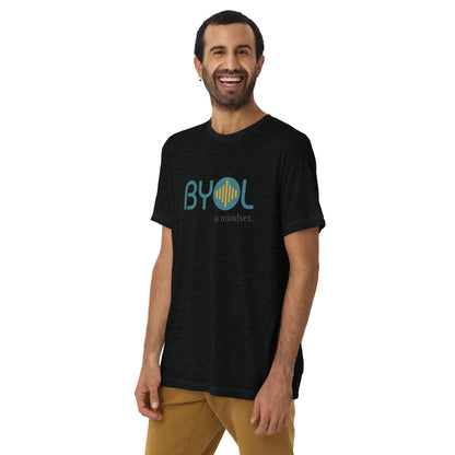 A young man with short dark hair and a beard wearing a black tri-blend "BYOL: a mindset" T-shirt, smiling and standing with his arms at his sides. The T-shirt features the "BYOL" logo in teal and is available in multiple colors (black, red, gray, pink, yellow, plus more) and sizes (S-3XL). The "be you out loud" logo is displayed at the top.