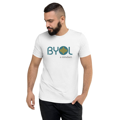 A young man with short dark hair and a beard wearing a solid white tri-blend "BYOL: a mindset" T-shirt, standing with one hand in his pocket and looking down. The T-shirt features the "BYOL" logo in blue and yellow on the front and is available in multiple colors (black, red, gray, pink, gold, plus more) and sizes (S-3XL). The "be you out loud" logo is displayed at the top.