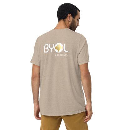 A young man with short dark hair wearing a sand tri-blend "BYOL: a mindset" T-shirt, viewed from the back. The T-shirt features the "BYOL" logo in white and yellow on the back and is available in multiple colors (black, red, gray, pink, beige, plus more) and sizes (S-3XL). The "be you out loud" logo is displayed at the top.