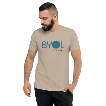 A young man with short dark hair and a beard wearing a tan tri-blend "BYOL: a mindset" T-shirt, standing with one hand in his pocket and looking down. The T-shirt features the "BYOL" logo in blue and yellow on the front and is available in multiple colors (black, red, gray, pink, gold, plus more) and sizes (S-3XL). The "be you out loud" logo is displayed at the top.