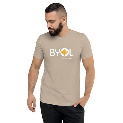 A young man with short dark hair and a beard wearing a heather sand "BYOL: a mindset" T-shirt, standing with one hand in his pocket and looking down. The T-shirt features the "BYOL" logo in white and yellow on the front and is available in multiple colors (black, red, gray, pink, gold, plus more) and sizes (S-3XL). The "be you out loud" logo is displayed at the top.