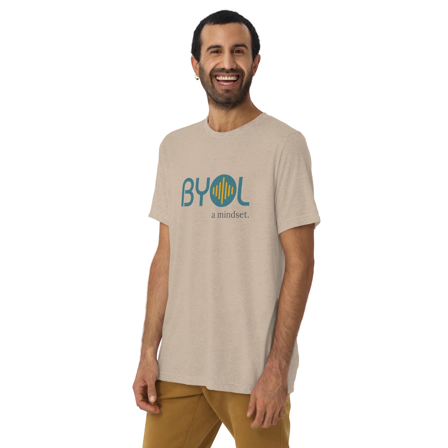 A young man with short dark hair and a beard wearing a sand tri-blend "BYOL: a mindset" T-shirt, smiling and standing with his arms at his sides. The T-shirt features the "BYOL" logo in teal and is available in multiple colors (black, red, gray, pink, yellow, plus more) and sizes (S-3XL). The "be you out loud" logo is displayed at the top.