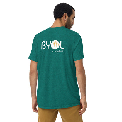 A young man with short dark hair wearing a teal tri-blend "BYOL: a mindset" T-shirt, viewed from the back. The T-shirt features the "BYOL" logo in white and yellow on the back and is available in multiple colors (black, red, gray, pink, beige, plus more) and sizes (S-3XL). The "be you out loud" logo is displayed at the top.