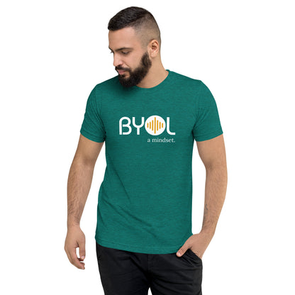 A young man with short dark hair and a beard wearing a emerald green tri-blend "BYOL: a mindset" T-shirt, standing with one hand in his pocket and looking down. The T-shirt features the "BYOL" logo in white and yellow on the front and is available in multiple colors (black, red, gray, pink, gold, plus more) and sizes (S-3XL). The "be you out loud" logo is displayed at the top.