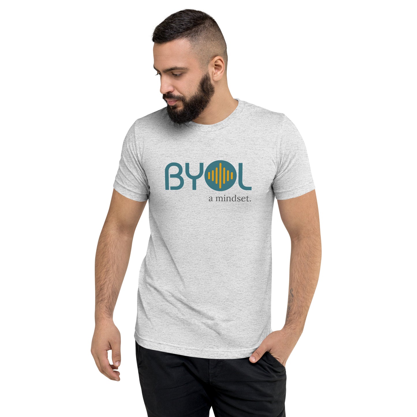 A young man with short dark hair and a beard wearing a white fleck tri-blend "BYOL: a mindset" T-shirt, standing with one hand in his pocket and looking down. The T-shirt features the "BYOL" logo in blue and yellow on the front and is available in multiple colors (black, red, gray, pink, gold, plus more) and sizes (S-3XL). The "be you out loud" logo is displayed at the top.