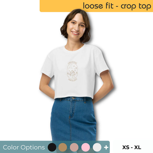 Woman wearing a white loose-fit crop top with a 'UNIVERSE UNFOLDER' celestial design, paired with a blue denim skirt, with size and color options available.