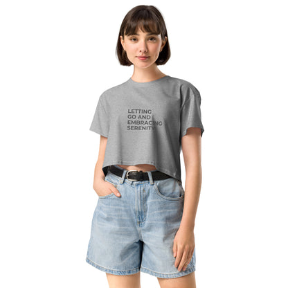 A young woman in a athletic grey cropped t-shirt with the text "Letting Go and Embracing Serenity" stands confidently with her hands in the pockets of her high-waisted denim shorts. She has a short bob haircut and is smiling slightly, creating a relaxed and serene vibe.