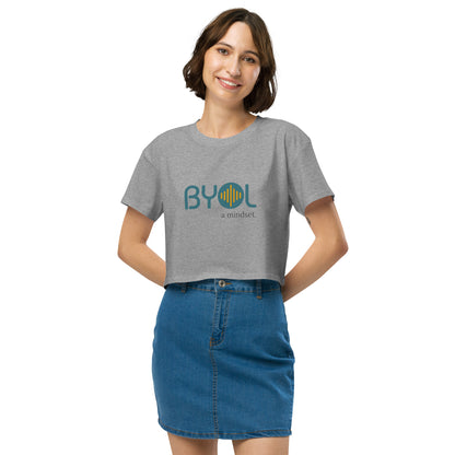 A young woman with short brown hair wearing a heather grey "BYOL: a mindset" crop top, smiling and standing with her hands on her hips. The crop top features the "BYOL" logo in teal and yellow on the front and is available in multiple colors (black, brown, white, light blue, plus more) and sizes (XS/SM - M/L). The "be you out loud" logo is displayed at the top.