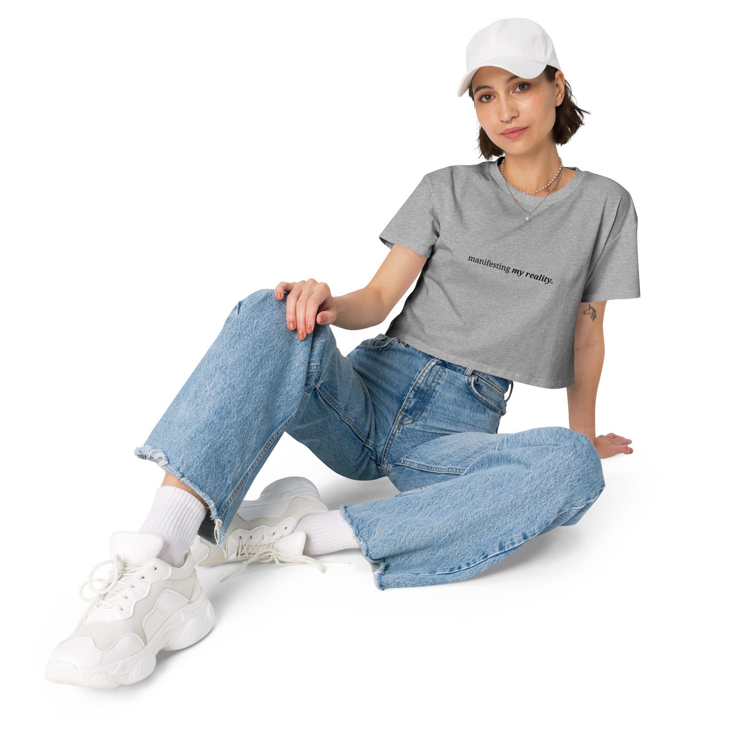 Woman wearing a heather grey cropped t-shirt with the phrase "manifesting my reality" printed in black, sitting and posing against a clean background. Available in various colors: black, brown, pink, green, and white. Sizes range from XS to XL.