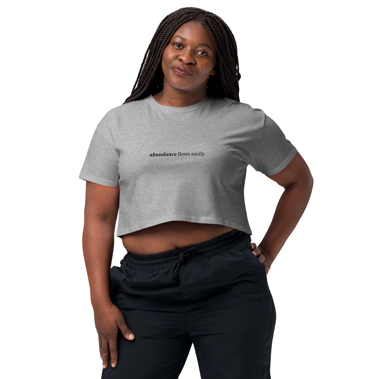 Woman wearing a heather gray cropped t-shirt with the phrase "abundance flows easily" printed in black, smiling and posing against a clean background. Available in various colors: black, brown, pink, green, and white. Sizes range from XS to XL.