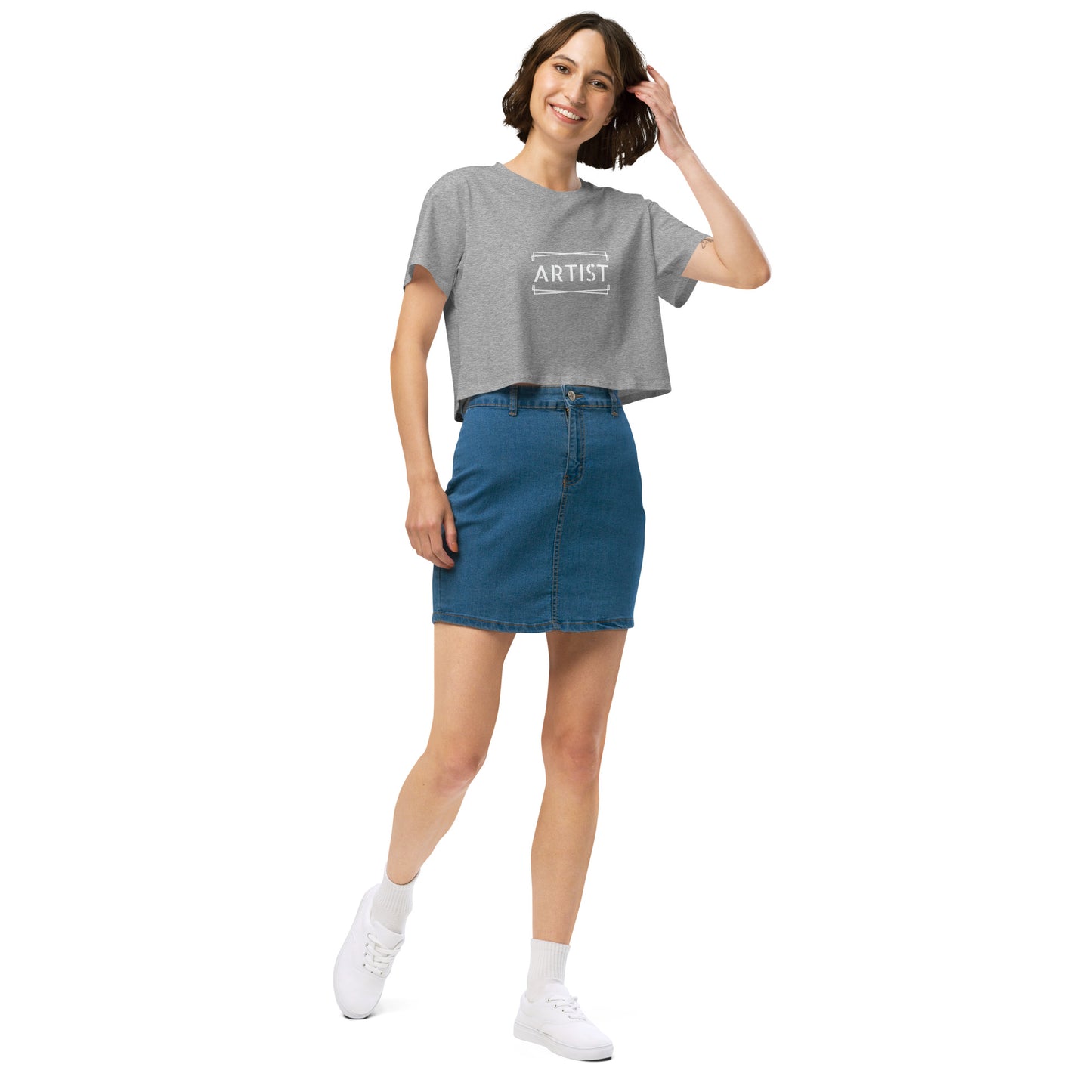 Stylish woman in a grey crop top emblazoned with 'ARTIST' in bold font, paired with a denim skirt, projecting a creative and expressive vibe.