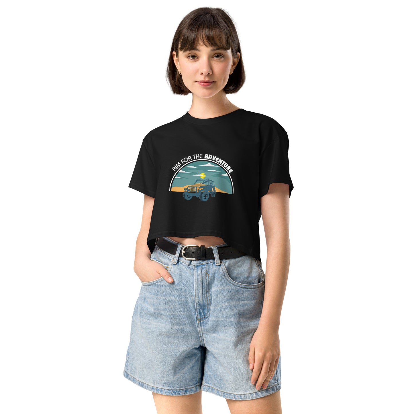 Content young woman wearing a black crop top with a vintage car and 'AIM FOR THE ADVENTURE' graphic, paired with light denim shorts for a laid-back, adventurous vibe.