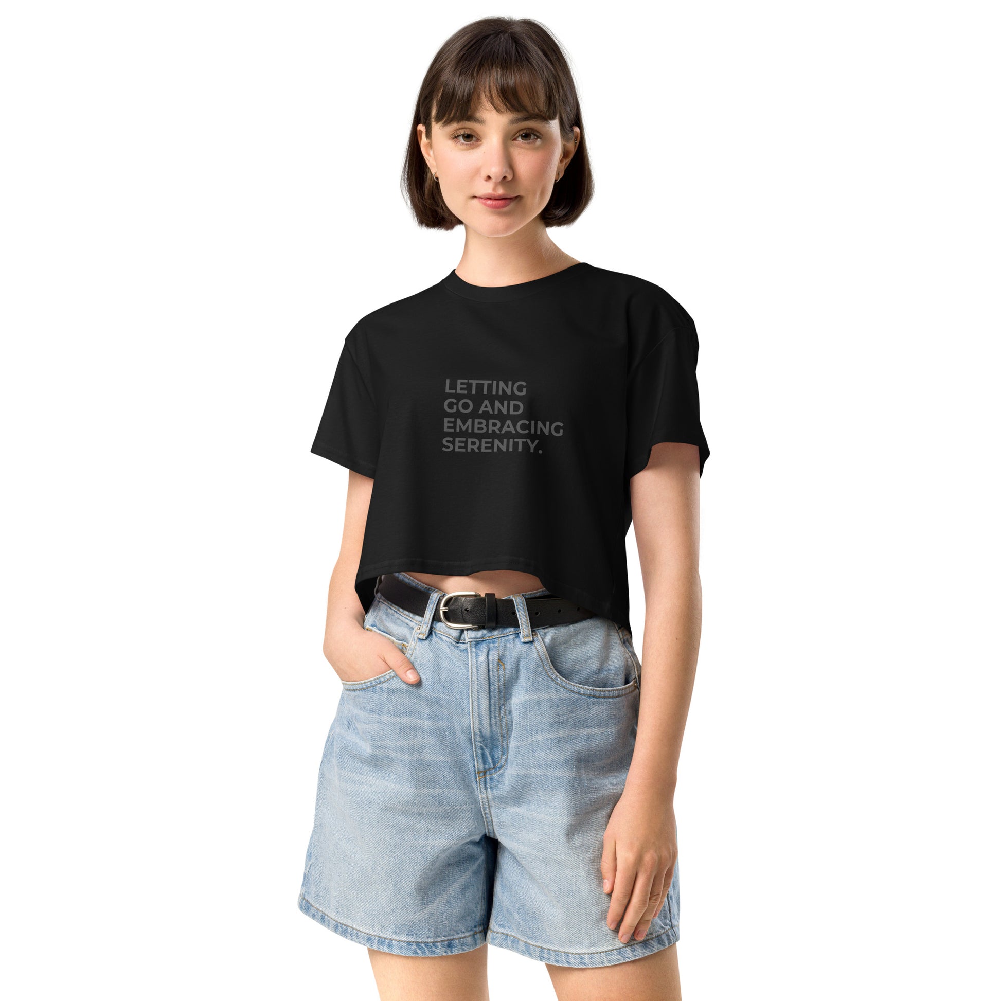 A young woman in a black cropped t-shirt with the text "Letting Go and Embracing Serenity" stands confidently with her hands in the pockets of her high-waisted denim shorts. She has a short bob haircut and is smiling slightly, creating a relaxed and serene vibe.