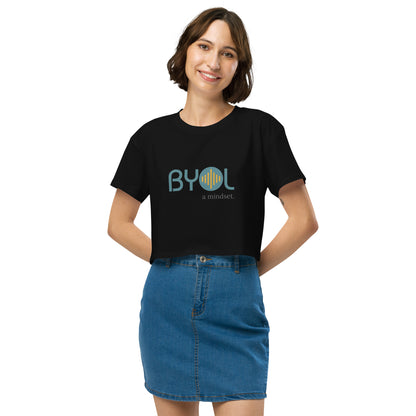 A young woman with short brown hair wearing a black "BYOL: a mindset" crop top, smiling and standing with her hands on her hips. The crop top features the "BYOL" logo in teal and yellow on the front and is available in multiple colors (black, brown, white, light blue, plus more) and sizes (XS/SM - M/L). The "be you out loud" logo is displayed at the top.