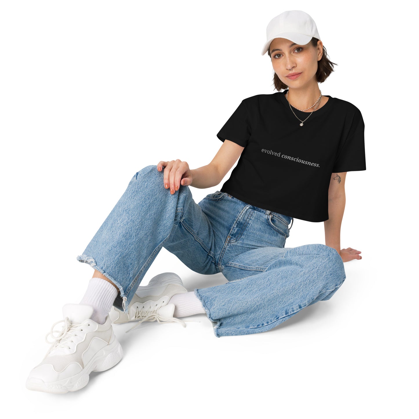 Woman wearing a black cropped t-shirt with the phrase "evolved consciousness" printed in black, sitting and posing against a clean background. Available in various colors: black, brown, pink, green, and white. Sizes range from XS to XL.