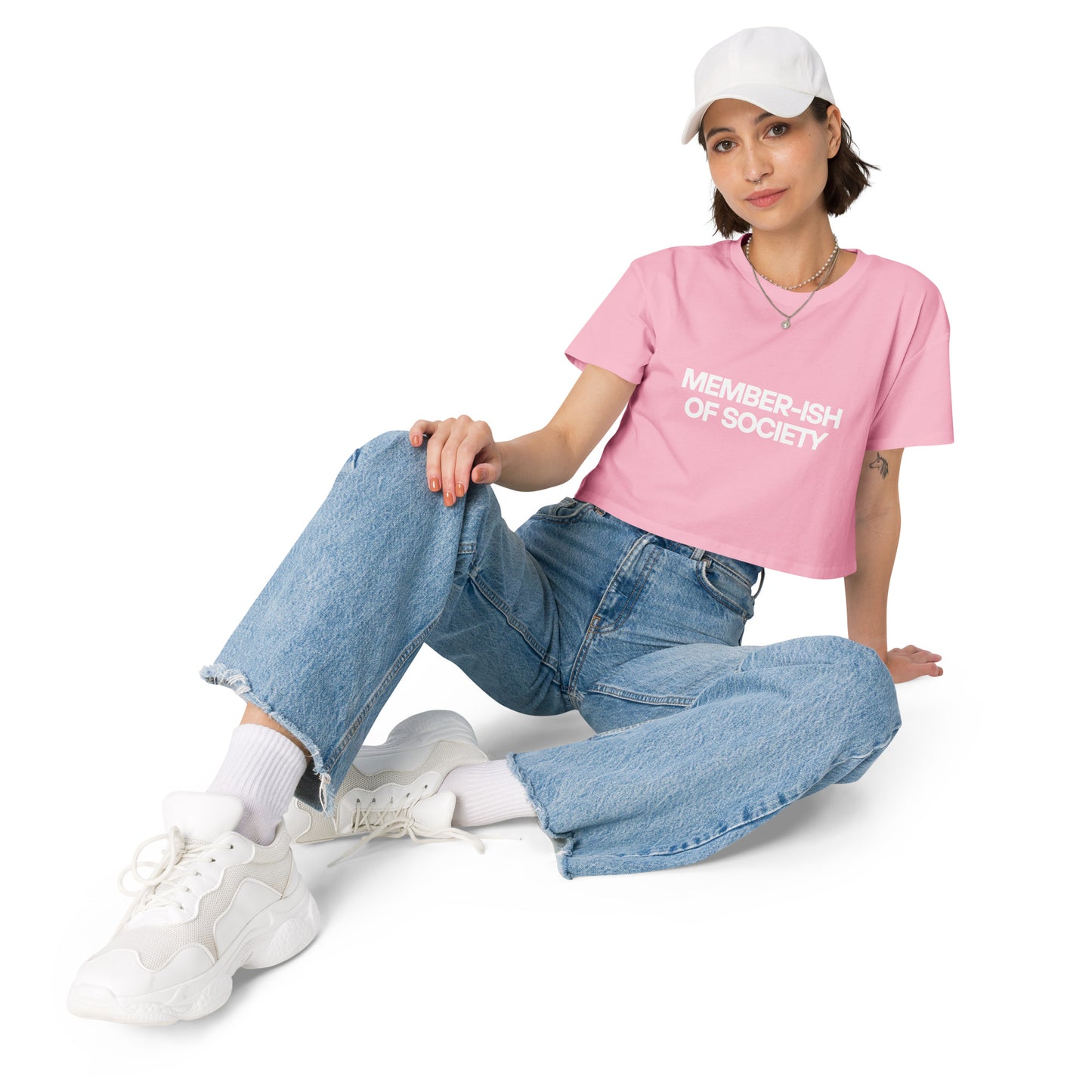 Casual and cool woman in a bubblegum crop top with 'Member-ish of Society' text, paired with denim jeans, white sneakers, and a cap for a laid-back look.