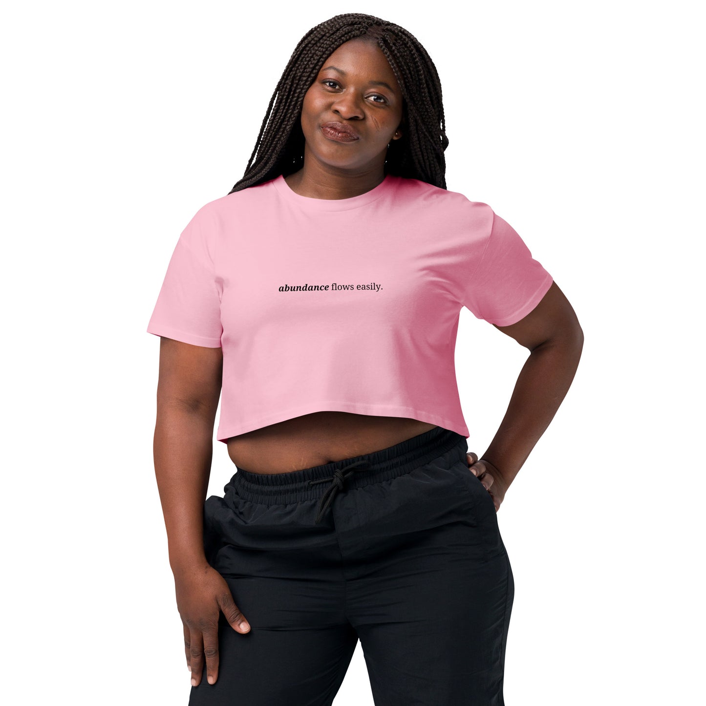 Woman wearing a bubblegum pink cropped t-shirt with the phrase "abundance flows easily" printed in black, smiling and posing against a clean background. Available in various colors: black, brown, pink, green, and white. Sizes range from XS to XL.