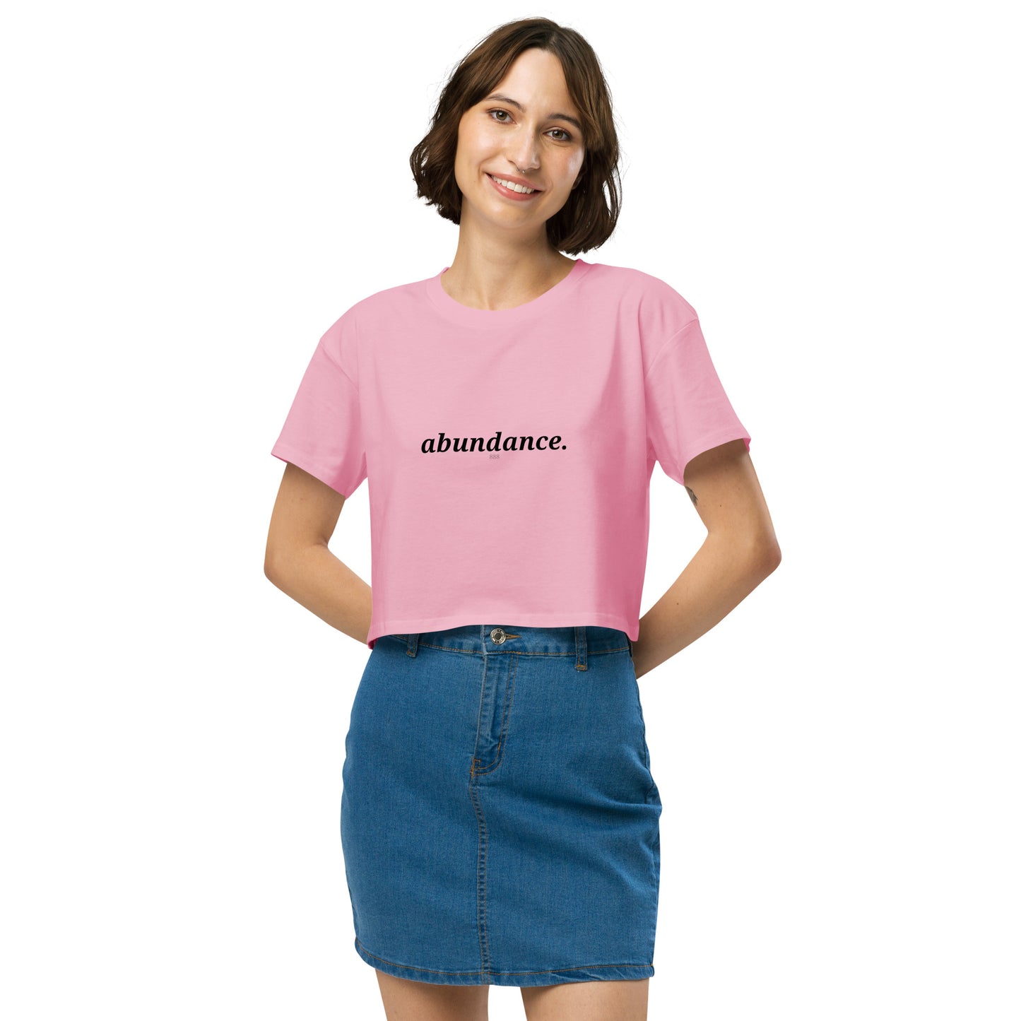 Woman wearing a bubblegum pink cropped t-shirt with the word "abundance" printed in black, smiling, and posing against a clean background. Available in various colors: black, brown, pink, green, and white. Sizes range from XS to XL.